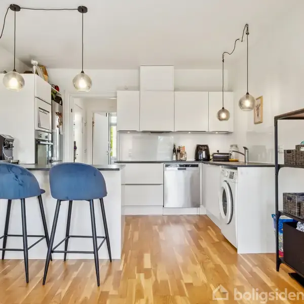 Et lyst køkken med hvide skabe og trægulv. To blå barstole står ved køkkenøen. Tre lamper hænger fra loftet. En vaskemaskine og opvaskemaskine er til højre.