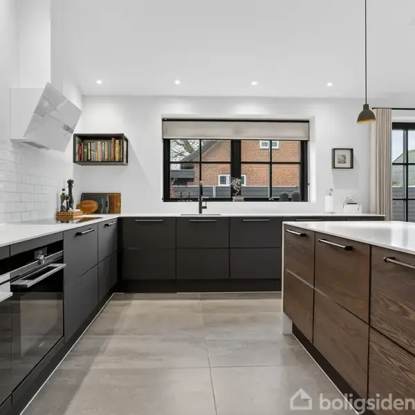Køkken med mørke skabe og hvide bordplader, indrettet med integrerede apparater, åbne hylder og en sort vask foran et stort vindue. Flisegulv og glat loft med indbyggede spots.