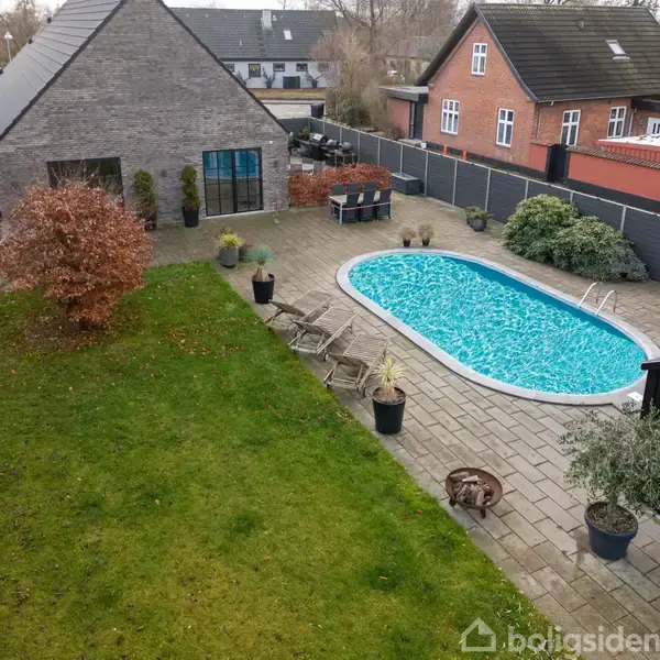 En udendørs pool ligger stille i en privat have med stenbelægning og græsplæne. Der er liggestole og potteplanter rundt om poolen, omgivet af murstenshuse og en hæk.