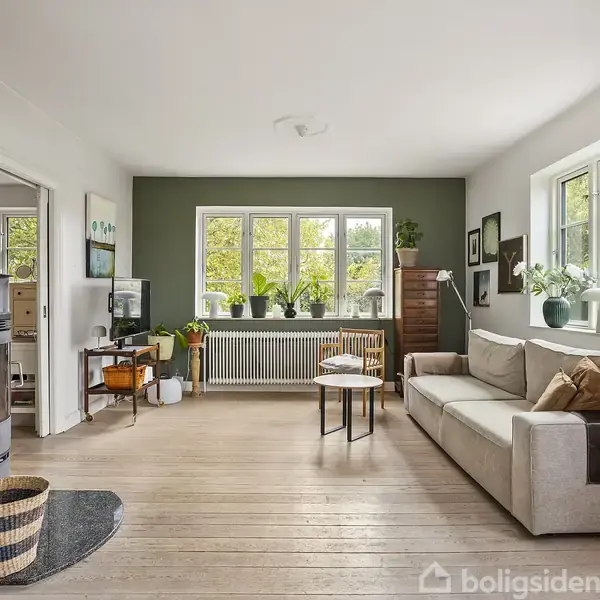 En lys stue med en beige sofa og træmøbler står i fokus. Rummet er dekoreret med planter og kunst på væggene. Store vinduer giver naturligt lys.