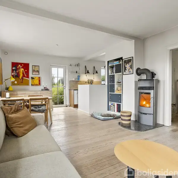 En lys stue med en grå sofa og brændeovn. Bord og stole ses ved vinduet. Væggene er dekoreret med kunst, og en åben dør fører til et andet rum.