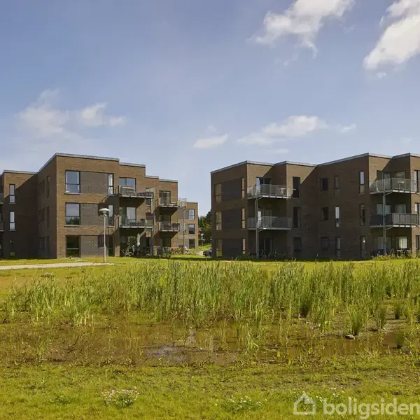 Flere moderne lejlighedskomplekser står stille, omgivet af grøn natur og en lille sø foran. Himlen er blå med spredte skyer.