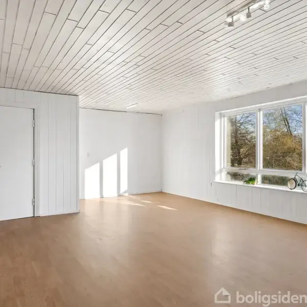 Et tomt, lyst værelse med hvidmalede vægge og trægulv. Et stort vindue til højre lader naturligt lys ind og viser træer udenfor. En lukket dør står til venstre.