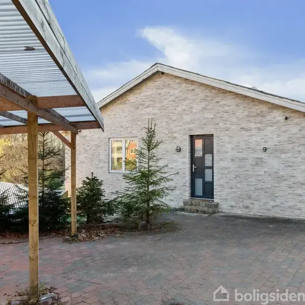 Huset står stille på en brolagt indkørsel omgivet af små grantræer. Det har en lys murstensfacade og blå dør. Til venstre ses en enkel carport med træstolper.