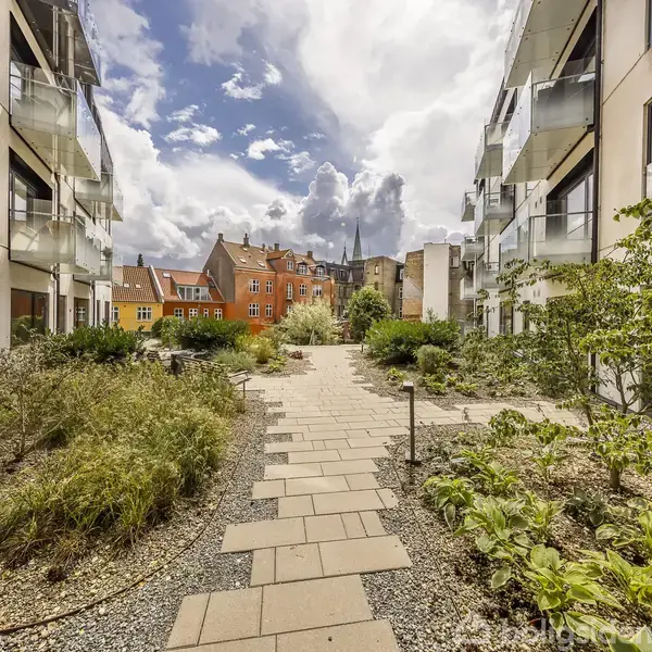 Et haveområde med gangsti omgivet af planter. Moderne lejlighedsbygninger ligger på begge sider i et bymiljø. Himlen er overskyet med sollys, der trænger igennem.