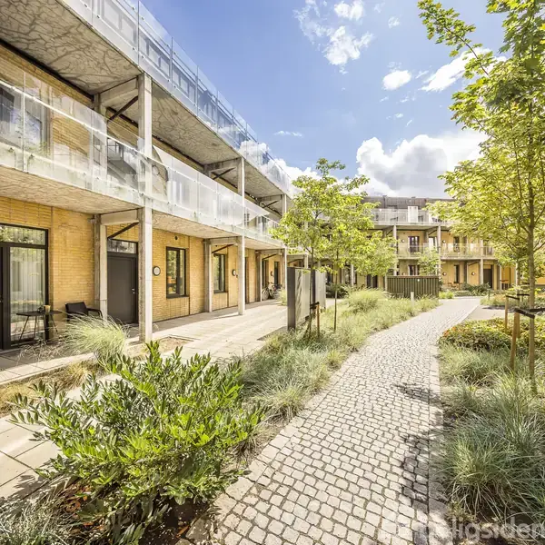 Gårdhave med brostensbelagt sti, omgivet af moderne gulstensbygninger og grønne planter under en blå himmel med skyer. Balkoner med glasværn skaber skygge og dybde i området.