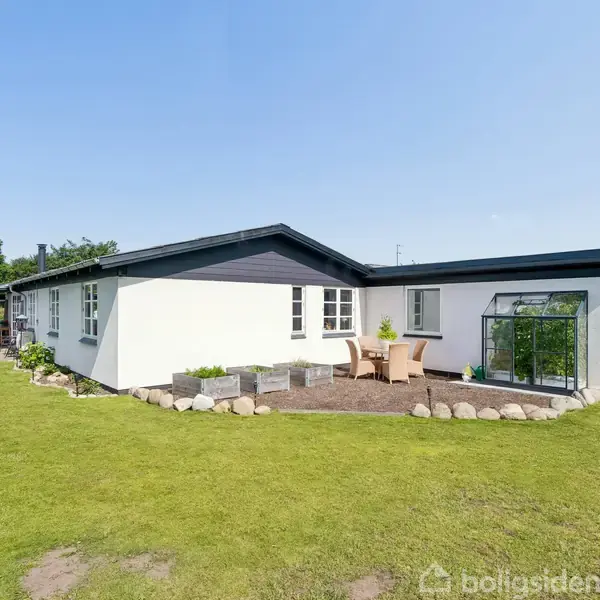 Et hvidt hus med sort tag står på en velplejet græsplæne. Rundt omkring er der blomsterkasser, et lille terrasseområde med borde og stole samt træer i baggrunden.
