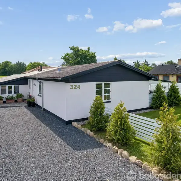 Et hvidt hus med nummer 324 står stille på en grusvej. Små buske flankerer en hvid stakit i en landlig omgivelse under en blå himmel.