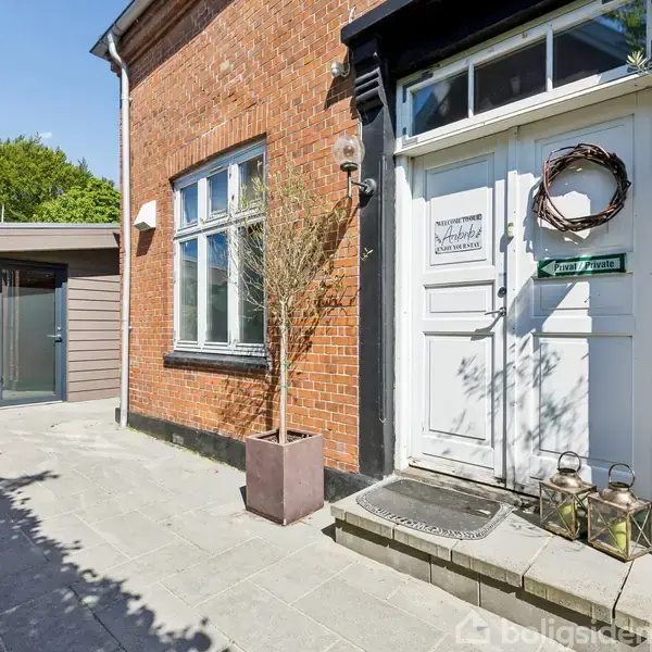 Hvid hoveddør med krans og lamper foran murstenshus, placeret på flisebelagt gårdhave. Til venstre ses et skur og grønne træer i baggrunden. Tekst på dør: "Drømmehuset."