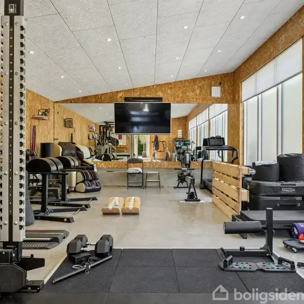Et fitnesscenter indeholder forskellige træningsmaskiner og vægtudstyr. Et stort spejl hænger på væggen foran en fladskærm. Store vinduer lader naturligt lys komme ind i rummet.