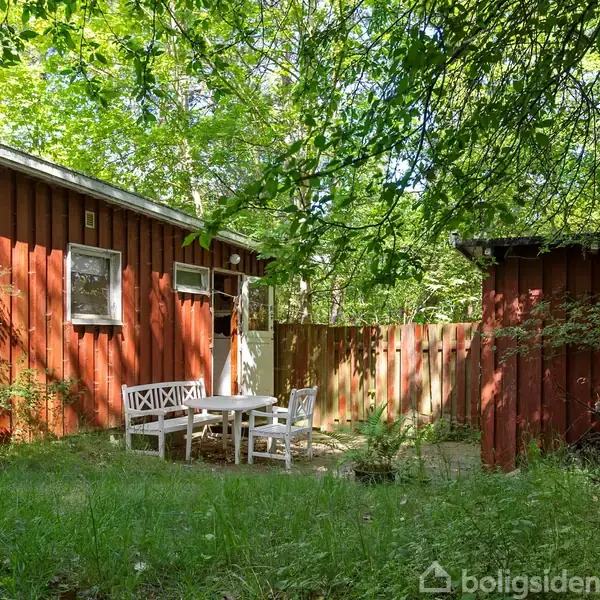 En rød træbygning står omgivet af tæt, grøn skov. Foran bygningen er der et hvidt siddesæt på en græsplæne, hvilket skaber en indbydende haveindretning.