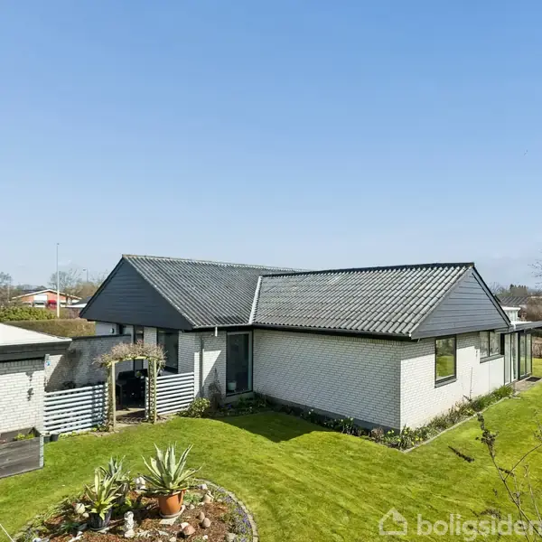 Huset står stille i en velholdt have med grønne plæner og få planter i forgrunden. Det har hvid mursten og et mørkt skråt tag, omgivet af en beboelsesområde.