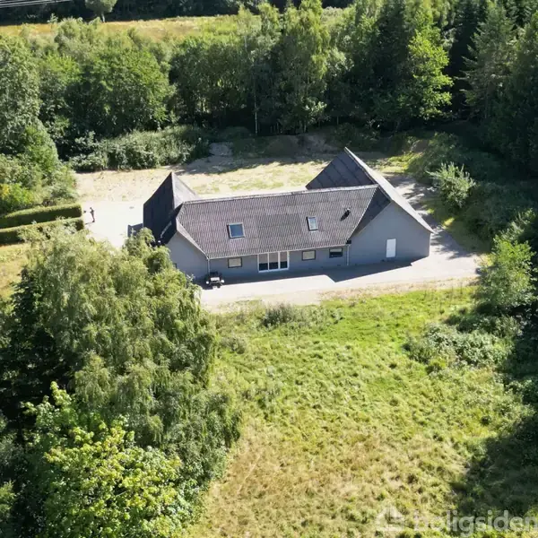 Grå bygning med skråt tag ligger omgivet af tæt skov og grønne marker. Sollys skinner på taget. Større åbning i skoven viser grusområde omkring huset.