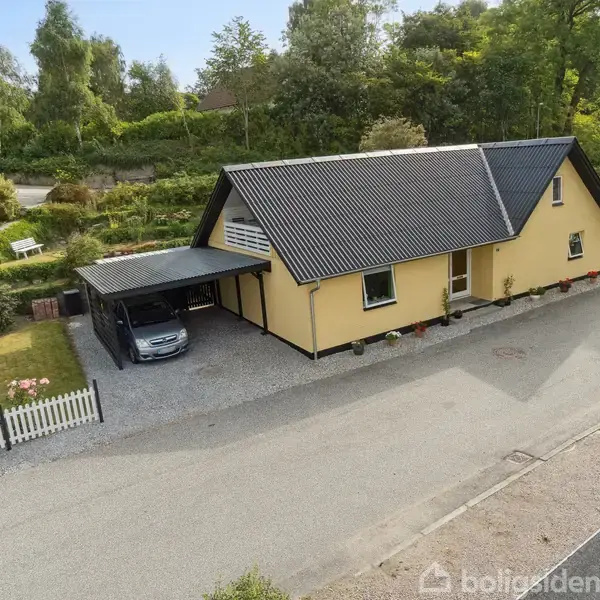 Et gult hus med sort tag står ved en stille vej. En bil er parkeret under en carport ved siden af huset, omgivet af en velplejet have og hegn.