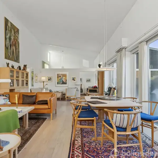 Stue med træmøbler og spisebord i forgrunden. Sofaer står ved væggen til venstre, pyntet med kunst. Store vinduer til højre for rummet giver lysindfald. Tæppe dækker gulvet.