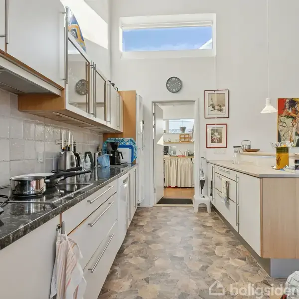 Køkken i moderne stil med lyse skabe og marmorbordplader, hvor der står forskellige køkkenapparater. På væggen hænger farverige billeder, og et ur er placeret over en døråbning.