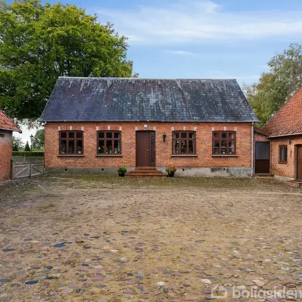 Et gårdsplads set med en central bygning af rød mursten og to fløje med tegltage. Bygningerne er omgivet af træer, og gårdspladsen har en brostensbelægning.