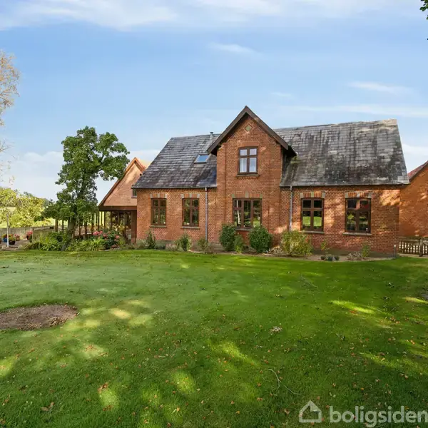 Rødt murstenshus ligger stille, omgivet af velplejet græsplæne og træer med blå himmel.