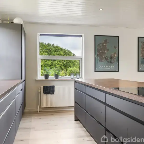 Et moderne køkken med mørkegrå skabe og en ø, dekoreret med krydderikrukker. To vægkort viser "DANMARK" og "SØRØHOLM". Lys trænges ind gennem vinduer med udsigt til grønne træer udenfor.