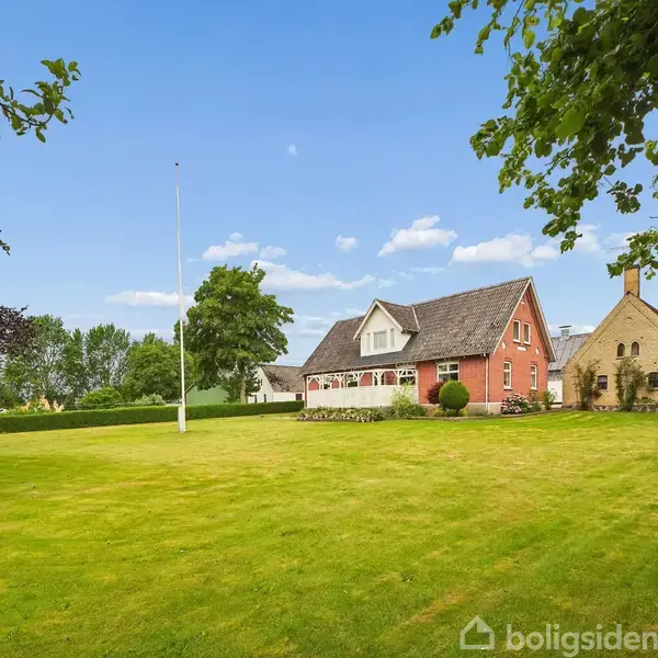 En stor rød villa står i en velholdt grøn have, omgivet af træer. En hvid flagstang er placeret til venstre for huset under en blå himmel med lette skyer.