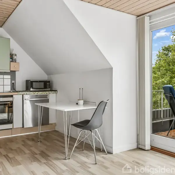 Et køkken med ovn, vaskemaskine og mikroovn nær en lille hvid bord med en stol. Glasdøre fører til en balkon med stole og udsigt over træer.