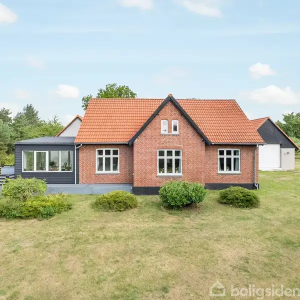 Rødt murstenshus med rødt tegltag står på en velplejet græsplæne omgivet af træer og buske. Der er en veranda på venstre side. Himmel med få skyer.