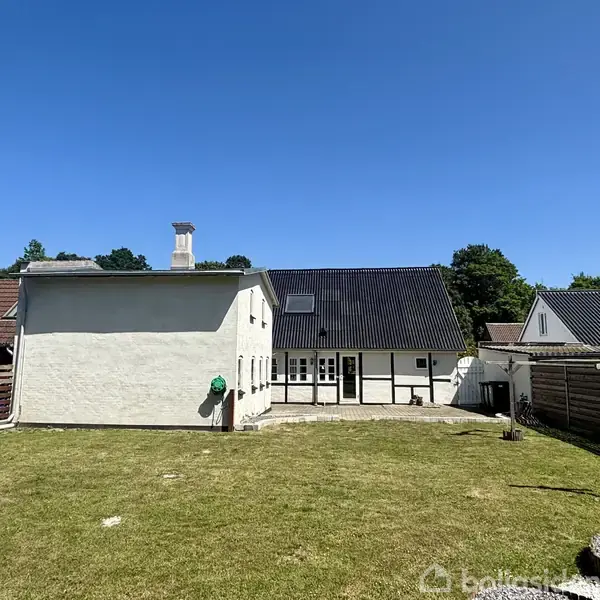 Et hvidt hus med sort tag står i en grøn have. Terrassen foran huset, omgivet af hegn, skaber en privat atmosfære. Klar blå himmel baggrunden.