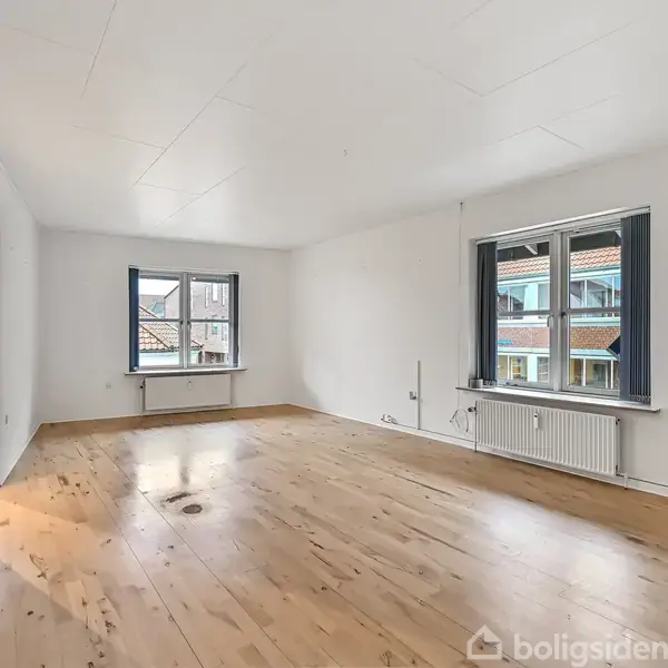 Et tomt rum med trægulv og hvide vægge. To vinduer åbner mod udendørs udsigt, og en radiator er placeret under det største vindue. En åben dør leder til et tilstødende rum.