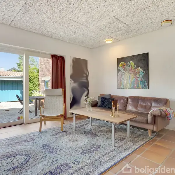 En stue med brun hjørnesofa ved siden af et sofabord. En stor, mønstret tæppe dækker gulvet. Der er glasdøre med udsigt til en terrasse og en blå bygning udenfor. To billeder hænger på væggen.