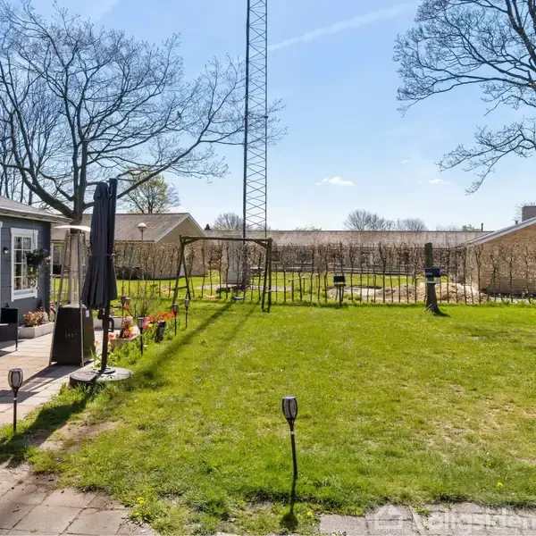 Terrasse med møbler står ved siden af en grøn græsplæne med en gynge. Omkringliggende træer og huse i baggrunden fuldender den fredelige haveatmosfære.