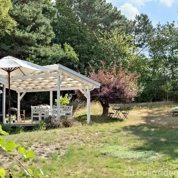 Blåt sommerhus under en pergola med hvidt cafébord og parasol, omgivet af frodig have med træer og to liggestole på græsset.