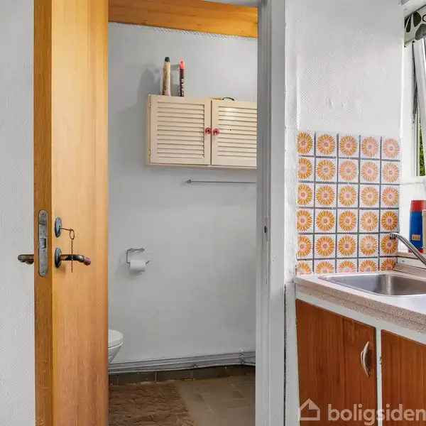 Dør åbner til et lille badeværelse med hvidt skab, toilet og vindue. Køkken til højre med vask, farverig flisevæg og udsigt til grønt udenfor.