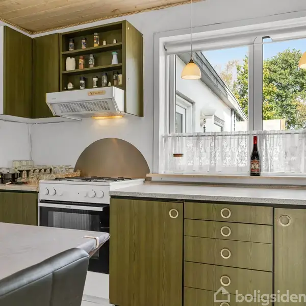 Et køkken med grønne skabe har en ovn med emhætte. Der står krydderiglas på hylden over ovnen. Et vindue med hvidt forhæng åbner ud til en have med træer.