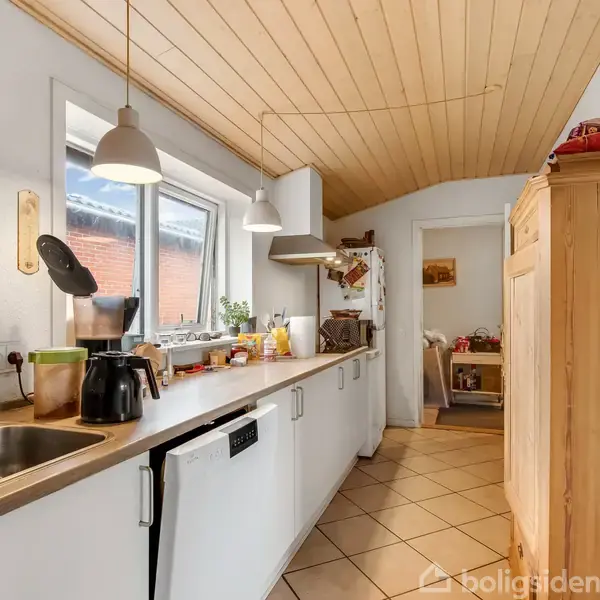 Køkken med lang bordplade fyldt med køkkenudstyr og planter placeret ved et vindue. En hvid opvaskemaskine står under bordpladen. Træloftet og skabe skaber en hyggelig atmosfære.