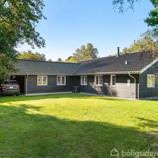 Hus står stille, omgivet af grøn græsplæne og træer, med carsport på venstre side. Sollys skinner på den mørke facade og hvide vinduer.