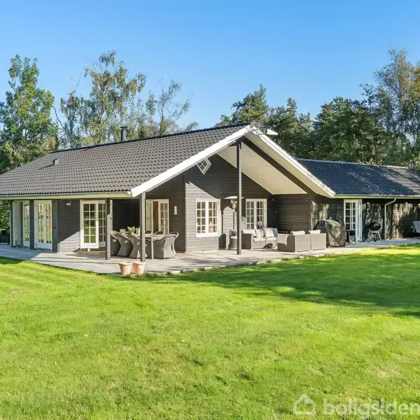 Et moderne sommerhus med sort tag står i en grøn have omkranset af træer. Der er en terrasse med havemøbler foran huset.