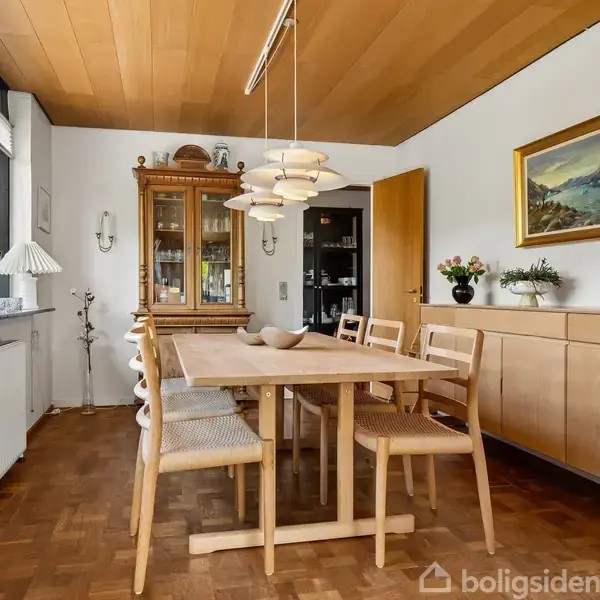 Træbord med seks stole står i en spisestue. En lampe hænger over bordet, omgivet af planter og vitrineskab. Billedet viser træloft og parketgulv.