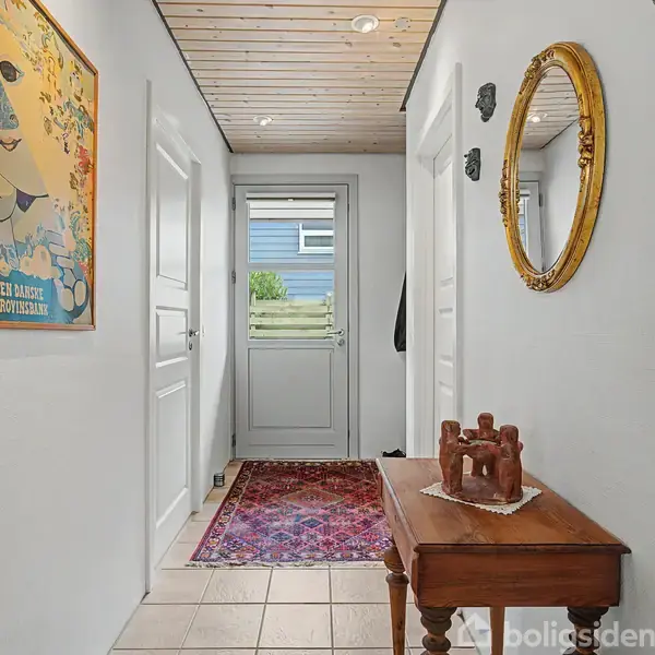 En gang med hvide vægge og loft, har en rød orientalsk tæppe på gulvet, en træskulptur på et bord, en rund guld-spejl på væggen og en dør mod en udendørs have. En plakat med teksten "EN DANSK EJERFORENING" hænger på venstre væg.