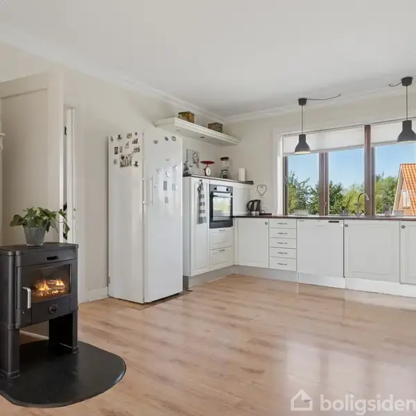En sort brændeovn opvarmer køkkenet, som har hvide skabe, hvidevarer og et stort vindue med udsigt til træer og hustage.
