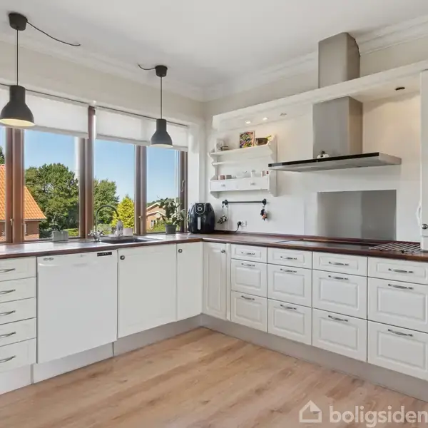 Et lyst køkken med hvide skabe, en indbygget kogeplade og emhætte. Store vinduer lader naturligt lys ind, og der er udsigt til et hus med rødt tag og træer udenfor.