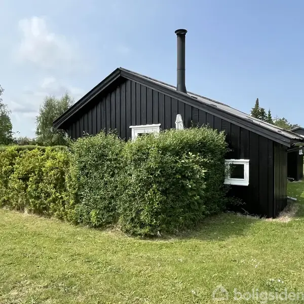 Sort træhus står stille, omgivet af grønne buske og græsplæne under en blå himmel. Het er en skorstenspibe på taget, omkranset af frodig natur.
