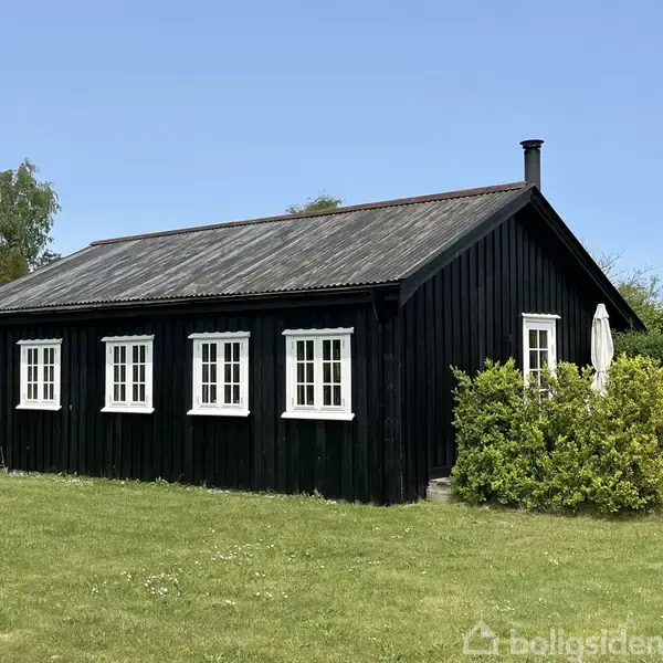 Et sort, træsommerhus med fem hvide vinduer står omgivet af grønne buske og græs. En hvid parasol er synlig ved siden af huset.