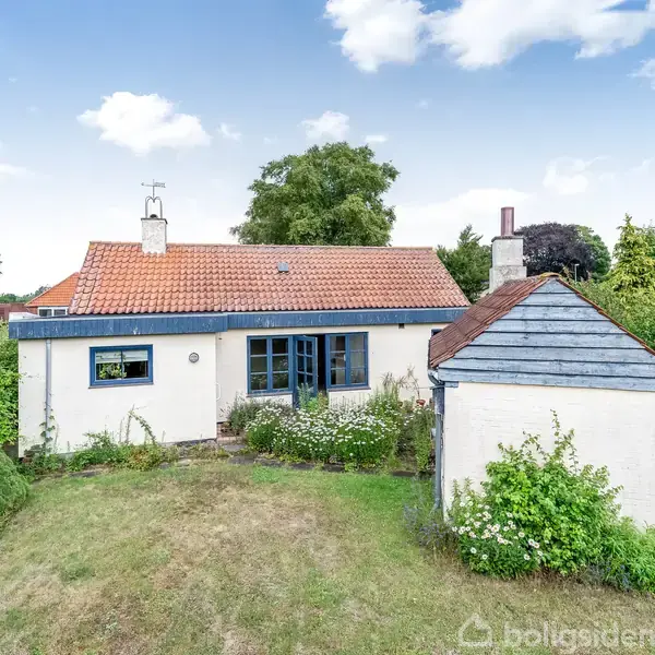 Et hvidt hus med rødt tegltag står i en frodig have med buske og græs, omgivet af andre huse og store træer under en blå himmel.