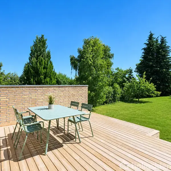 En terrasse med et bord og seks stole står stille i solskin. Terrasse af træ omgivet af en lav mur, med grønne træer og græsplæne i baggrunden under en klar blå himmel.