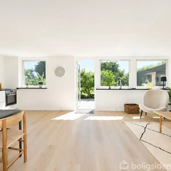 Et lyst, moderne køkken-alrum med træmøbler og lyse trægulve. Store vinduer og glasdør giver udsigt til en grøn have. En grå sofa og enkel indretning fuldender rummet.