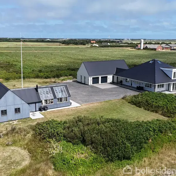 Én moderne bygning med mørkt tag og tilstødende garage står nær et lille drivhus, placeret på en åben mark med udsigt over flade marker og få spredte bygninger i horisonten.
