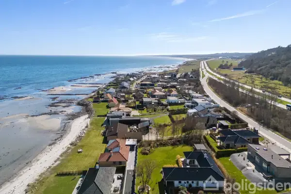 Kystby med huse langs en strand. Husene strækker sig parallelt med en vej, der løber langs kysten. Langt horisonten ses havet, og området er omgivet af grønne bakker.