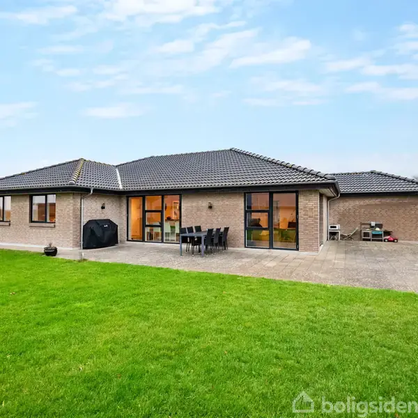 Et moderne, én-etagers hus med fladt tag står stille omgivet af en velplejet græsplæne og terrasse med havemøbler. Sollys skinner gennem store vinduespartier til venstre og højre.