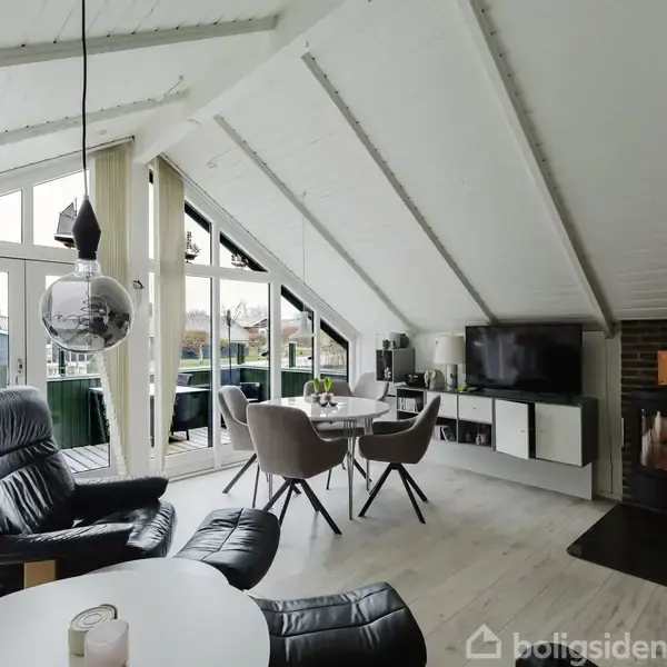 Stue og spiseområde under skrå loft med store vinduer, lænestole i sort læder, rundt spisebord og brændeovn i hjørnet; lyse nuancer dominerer interiøret.