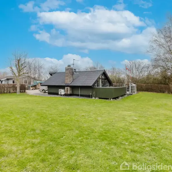 Et sort træhus står stille midt på en stor, grøn græsplæne, omgivet af nøgne træer og en hæk. Himlen er blå med spredte hvide skyer.